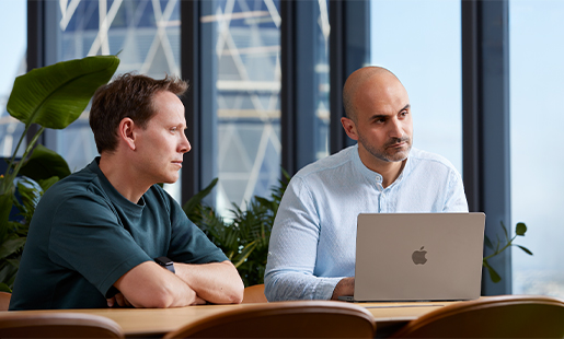 Ahmed est assis à une table avec son MacBook devant lui, à côté d’un collègue. Il regarde une personne qui n’est pas dans le cadre.