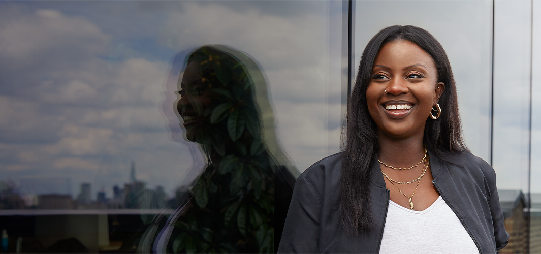 Ginelle, à l’extérieur d’un bâtiment, regarde au loin vers la droite en souriant.