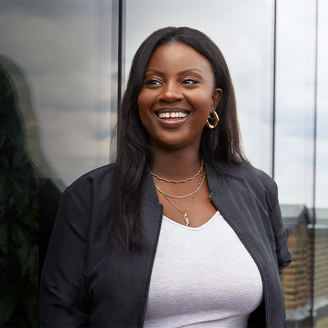 Ginelle, à l’extérieur d’un bâtiment, regarde au loin vers la droite en souriant.