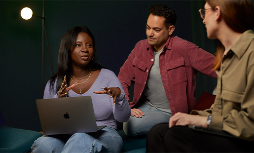 Ginelle est assise avec un MacBook sur les genoux et travaille avec deux collègues.