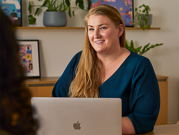 Une employée Apple est assise à une table dans un espace collectif. Elle utilise un MacBook et discute avec une collègue.