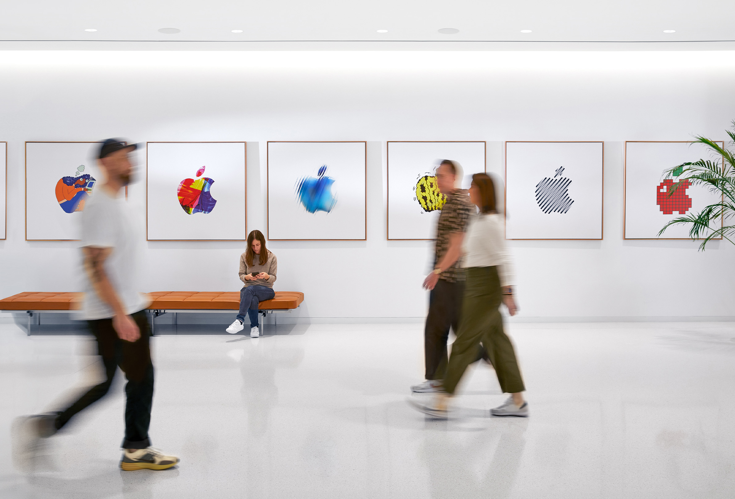 Trois membres de l’équipe Apple marchent devant une cloison décorée de logos Apple colorés. Une quatrième employée est assise sur un banc.