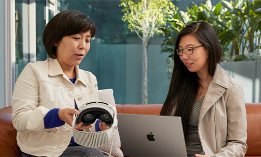 Jessica utilise un MacBook dans un espace intérieur, devant une baie vitrée qui donne sur une terrasse. Elle est assise avec une collègue Apple qui lui montre l’Apple Vision Pro.