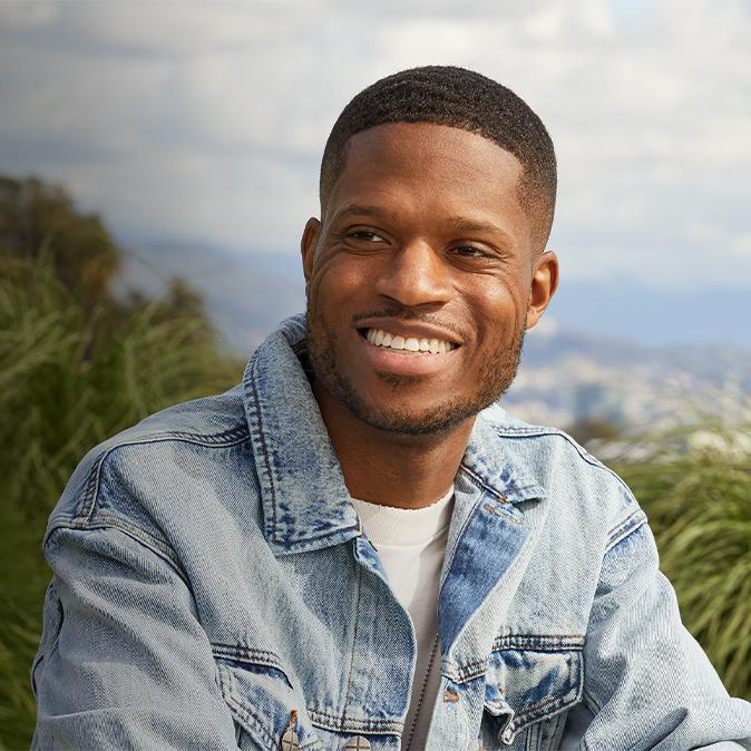 Justin est assis sur une terrasse extérieure, souriant. Le ciel et des arbres se trouvent en arrière-plan.