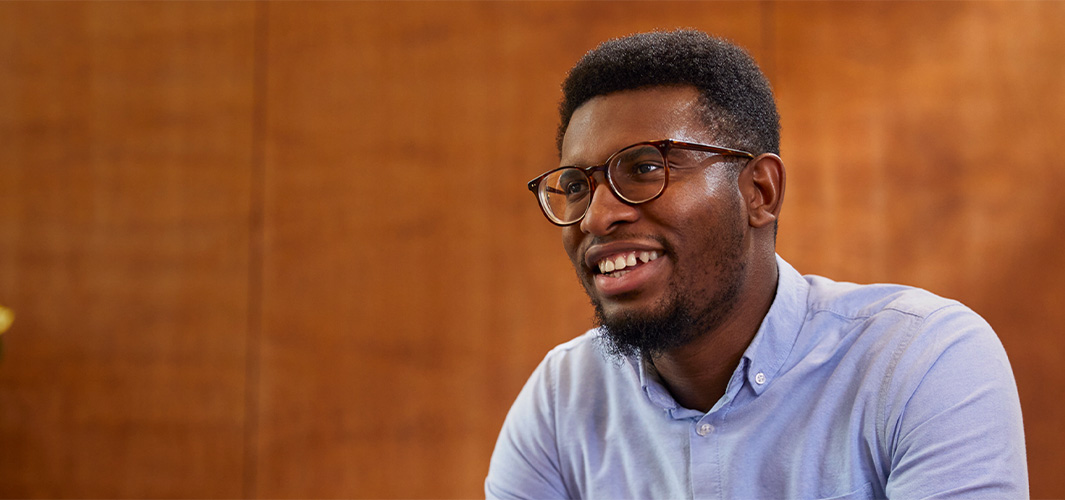 Chidi, assis, sourit, le regard tourné sur le côté.