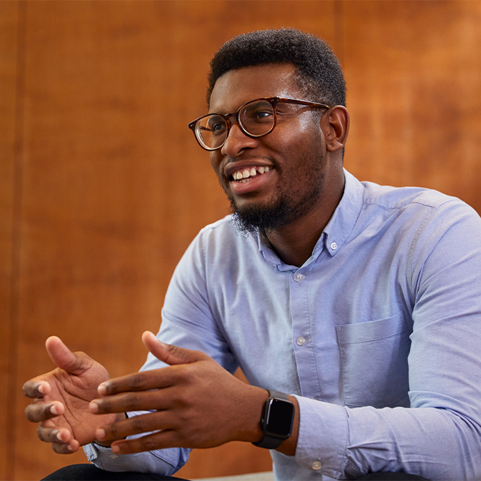 Chidi, assis, sourit, le regard tourné sur le côté.