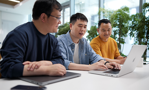 Ken écrit sur son MacBook, un membre de l’équipe assis à sa gauche et à sa droite, et un arrière-plan composé de fenêtres lumineuses et de plantes.