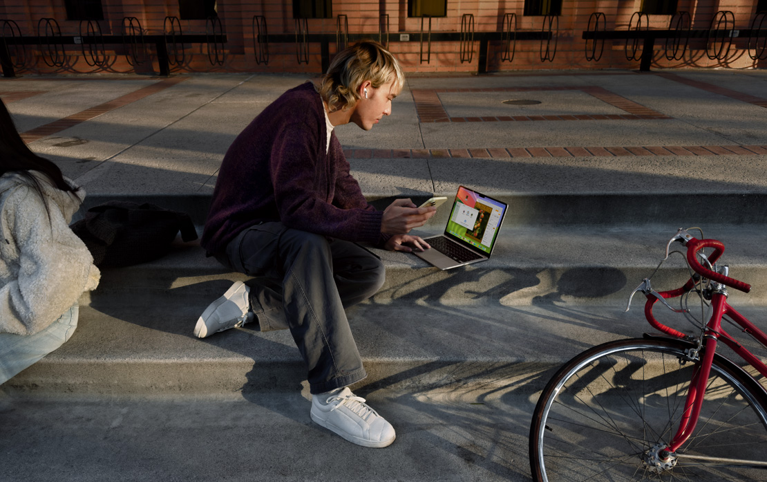 Un estudiante universitario sentado en unas escaleras usa un iPhone y un MacBook. A su lado, una bicicleta.