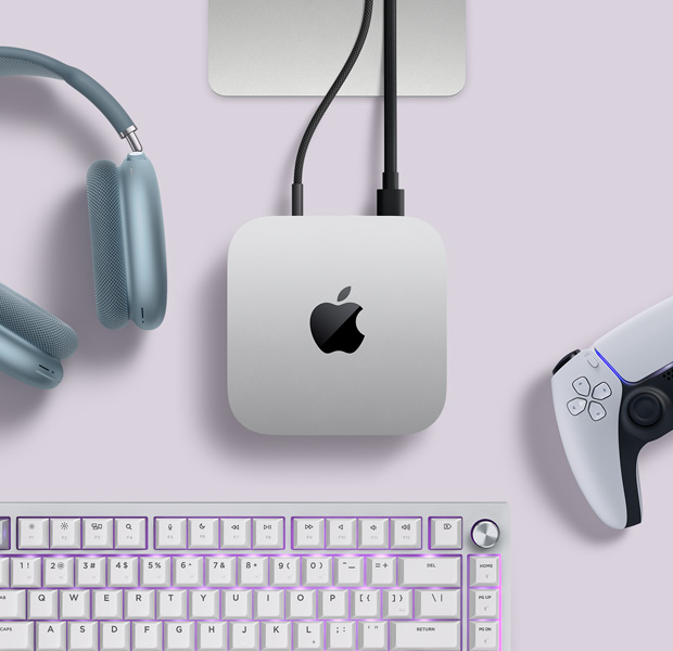 Imagem de cima do Mac mini prateado, com dois cabos conectados às portas de trás, ao lado de AirPods Max azuis, um Magic Keyboard prateado e um controle sem fio para jogos.