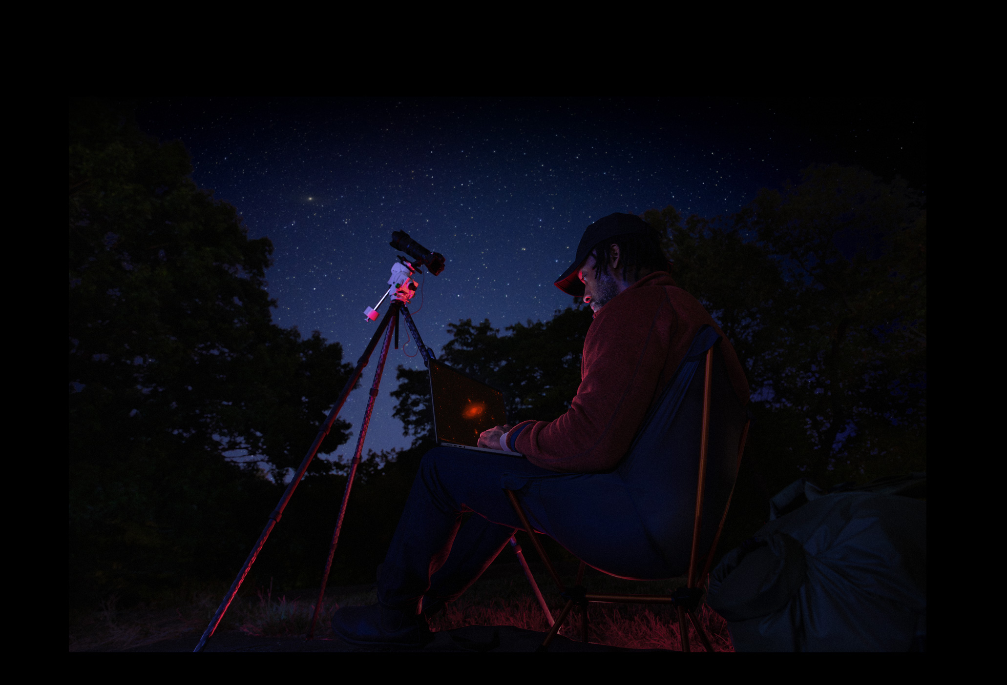 Una persona que está al aire libre, de noche, tiene un MacBook Pro apoyado en su regazo conectado a un dispositivo con un trípode que apunta hacia las estrellas